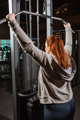 overweight girl in grey hoodie doing arms extension exercise on fitness machine