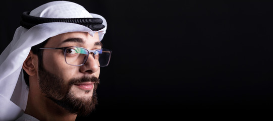 Portrait of handsome young Arabic man with glasses and white agal on dark background.