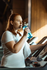 Naklejka na ściany i meble pensive overweight girl drinking from sports bottle while standing at treadmill