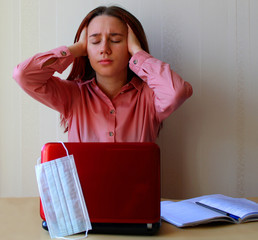 A girl sits clutching her head and closing her eyes at the table with a computer and a copybooks. Distance education in universities and institutes due to the epidemic of coronavirus Covid-19