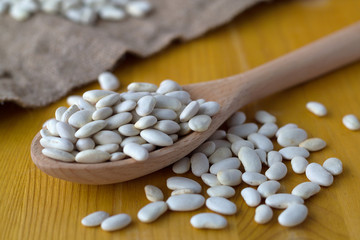 close-up white beans in a wooden spoon on a yellow background. uncooked white beans with sackcloth on a wooden table. Healthy lifestyle.
