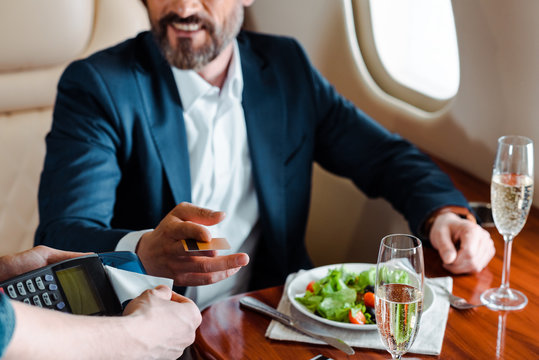 Cropped view of man holding terminal near smiling businessman with credit card, salad and champagne on table in plane
