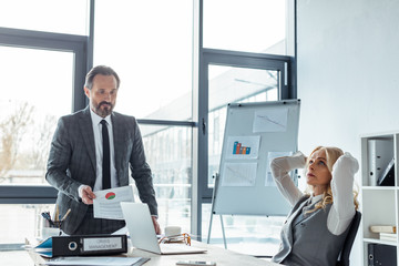 Selective focus of thoughtful businesswoman sitting at table near businessman holding paper with chart in office