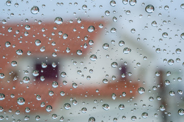 Texture of water drops on window glass on a sad rainy day. Natural pattern of raindrops on Mediterranean style tiled roof houses background. Raining spring on coast. Reflection of the house in drops.
