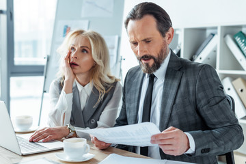 Handsome businessman holding document near thoughtful businesswoman using laptop at table in office