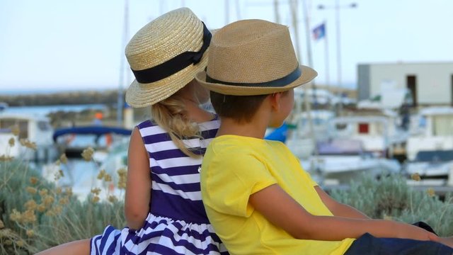 Little boy and adorable blond girl in straw hats are sitting with theirs back to the camera on the background of the yacht port