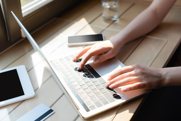 cropped view of woman typing on laptop keyboard near credit card and gadgets with blank screen