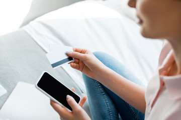 selective focus of woman holding smartphone with blank screen and credit cards in bedroom