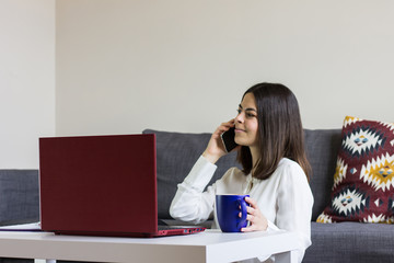 Happy casual beautiful woman working on a laptop sitting on the floor at home with cup of coffee in hand and talking on the phone.