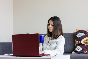 Happy casual beautiful woman working on a laptop sitting on the floor at home with cup of coffee in hand.