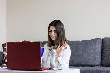 Happy casual beautiful woman working on a laptop sitting on the floor at home with cup of coffee in hand and saying hello.