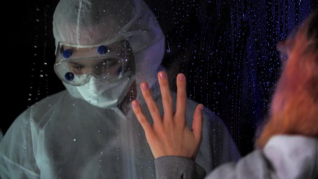 Man in hazmat protective medical suit shows indecent gesture, middle finge to sad woman, who touch palms to glass through the window to the hospital. Quarantine or self isolation