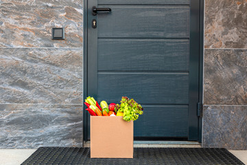 Contactless delivery during a pandemic flu. A box of fresh vegetables and fruits from a supermarket stands at the front door. Stay home. Coronavirus. Quarantine.