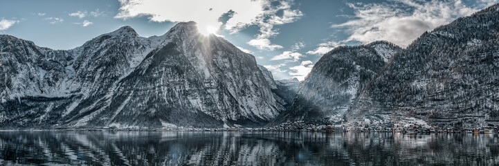Panoramic view of snowy Hallstatt village houses at foot of mountain by lake with a ray of sunshine in winter in Austria