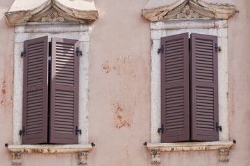 Fototapeta na wymiar Two Italian windows on the pink flaky wall facade with closed wooden brown color classic shutters