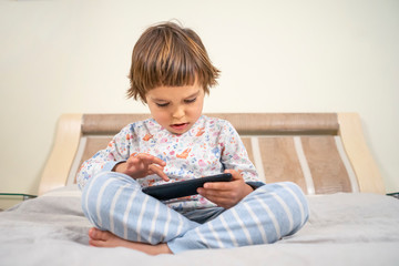 Fototapeta premium A little boy plays games on the phone. A child sits on a bed in pajamas with a phone