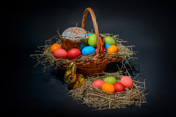 Easter colored eggs and Easter cake with straw in wicker basket on black background