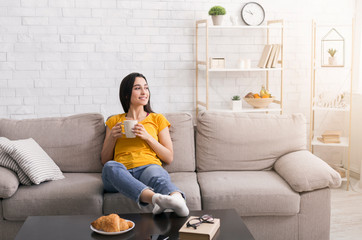 Stay home, drink coffee. Millennial girl with cup of hot beverage relaxing on comfy sofa in living...