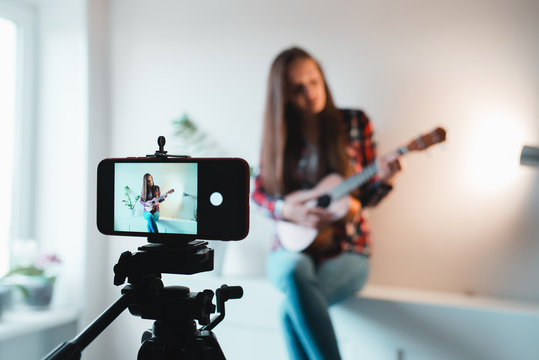 Girl in a shirt and jeans writes a vlog on the phone about playing ukulele.