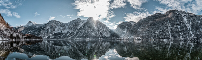 Fototapeta premium Panoramic view of snowy Hallstatt village houses at foot of mountain by lake with reflection in winter in Austria
