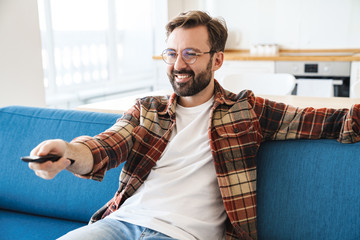 Portrait of young bearded man smiling and watching television at home
