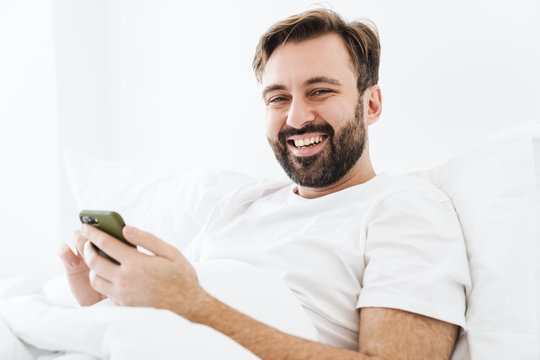 Image of young unshaven caucasian man smiling and using cellphone while lying in bed at home