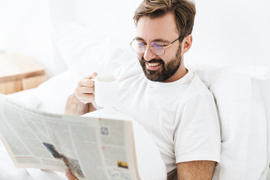Image of young man drinking coffee and reading newspaper in bed at home