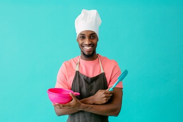 Portrait of a smiling young male chef with arms crossed holding spatula and bowl, against blue...