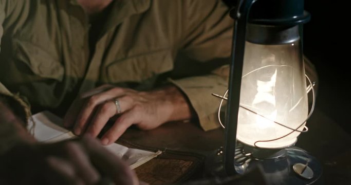 Military man writing strategy of defeating enemy. Tilt up of World War II soldier sitting at table with burning kerosene lamp and taking notes of strategy while sitting in enemy rear