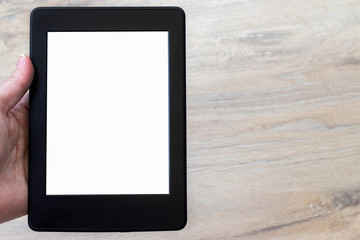 A modern black electronic book with a white blank empty screen in human hand against a blurred wooden floor background. Mockup tablet closeup with copy space