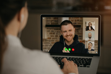 Fototapeta premium Back view of a woman talking with a business partner and colleagues in a video call on a laptop. Man talks with coworkers on a webcam conference. Multiethnic business team having an online meeting