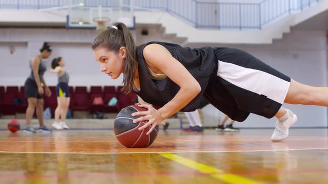 Young female athlete doing push ups with basketball and then sticking out her tongue and falling down on court while getting tired after workout
