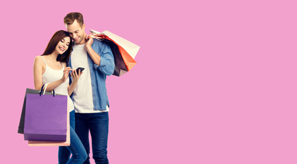 Holiday sales, online shopping, consumer concept - happy couple with bags, looking at mobile phone, standing close to each other. Rose pink color background. Copy space for some text.