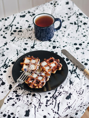 An excellent early Breakfast. Viennese Belgian waffles decorated with flowers on a black plate, a mug of black tea and vintage Cutlery on the table. White background and black and white tablecloth