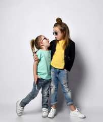 Two frolic kid girls sisters in stylish casual clothing, sunglasses and sneakers stand together and...