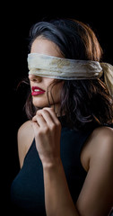 Portrait of beautiful young woman tying her eyes with white scarf on black background
