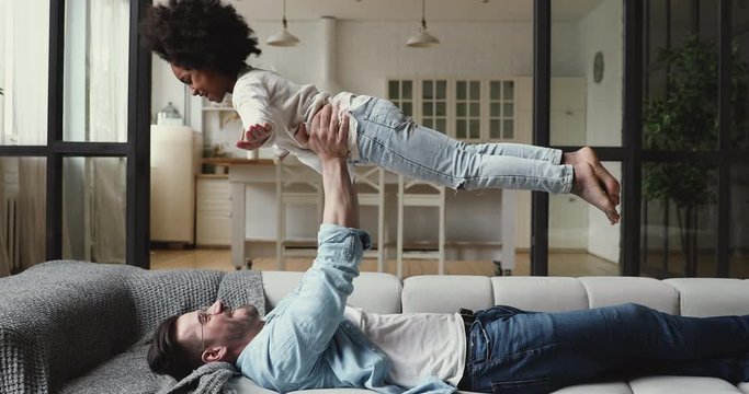 Happy caucasian parent stepfather playing plane game lifting cute small african kid daughter relaxing on couch. Cheerful mixed ethnicity family young dad and child girl having fun together at home.