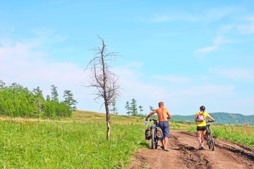 Two cyclist, man and woman, biking on a bike lane inside a park with flower field and blue sky.