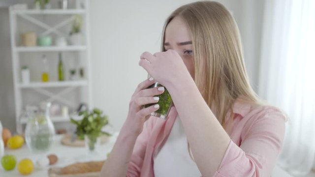 Unhappy Caucasian girl closing nose with fingers to drink green healthful cocktail. Portrait of slender young blond woman with healthy drink indoors. Lifestyle, health care, dieting.