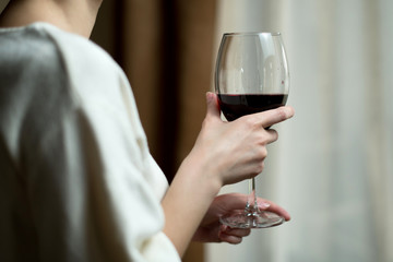 Girl holds a glass with red wine, standing near the curtained window.