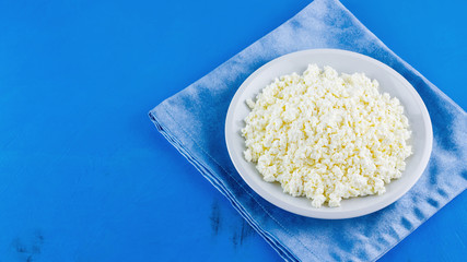 Cottage cheese in a white plate on a blue background. Organic eco healthy meal, dairy product. Plate of cottage cheese on a blue napkin. Top view. Copy space