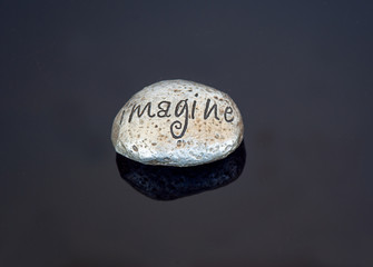 Painted rock imagine word on a shiny black background.  The rock is a metallic silver color, great for concepts and ideas for creativity and imagination.