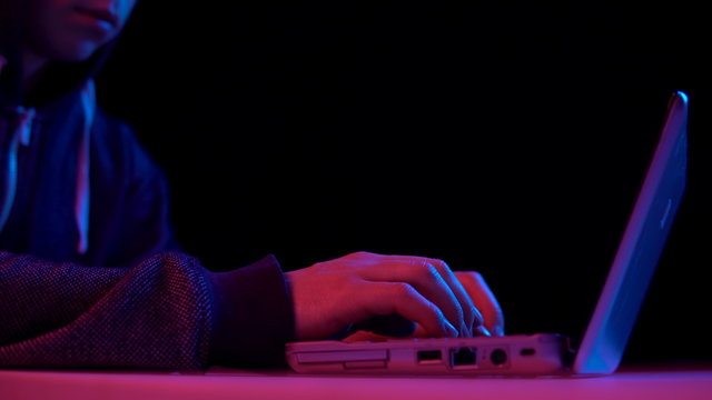 Young man in a hood with a laptop closeup. Hacker makes a hack through a laptop. Blue and red light falls on a man on a black background.