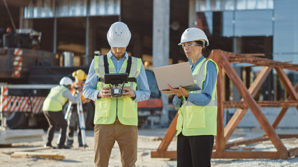 Two Specialists Controlling Drone on a Construction Site. Architectural Engineer and Safety Engineering Inspector Fly Drone on Commercial Building Construction Site Controlling Design and Quality