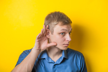 Young, serious, blond man in a blue shirt on a yellow background, holding an ear to listen better,...