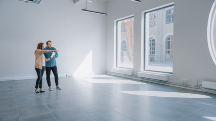 Fototapeta premium Young Hipster Man and Female Stand in an Empty White Office and Map it with an Augmented Reality Software on a Tablet. Sunlight Shines Through Big Windows.