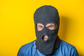 man close up thief in a mask and a blue shirt on a yellow background looks slyly to the camera....