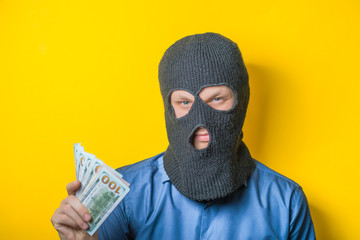 man close up thief in a mask and a blue shirt on a yellow background looks slyly to the camera....
