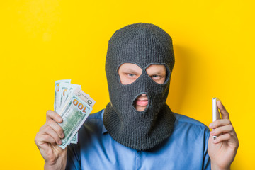 man close up thief in a mask and a blue shirt on a yellow background looking slyly at the camera, holding the money and the phone. Mimicry. Gesture. photo Shoot
