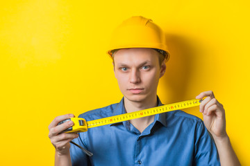 Young man close-up in a blue shirt and construction helmet on a yellow background holding a tape measure, measure something. Mimicry. Gesture. photo Shoot